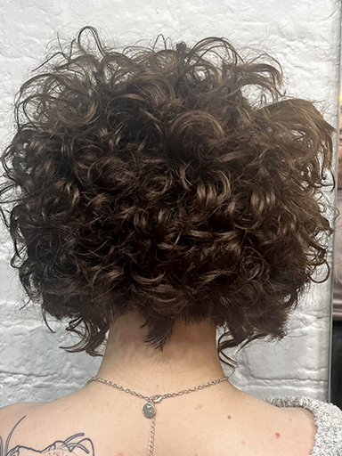 Curly brown hair styled voluminously on a person with a tattooed shoulder, set against a textured white wall, creating a lively and dynamic look.