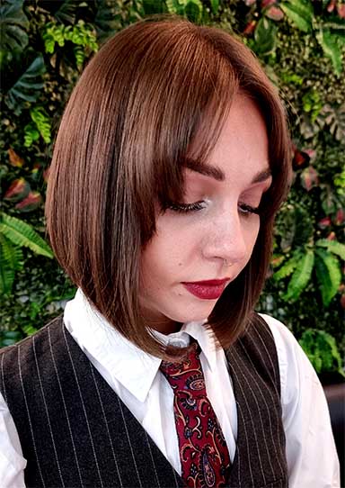A person with a bob haircut, wearing a pinstripe vest, white shirt, and red patterned tie, looks downwards. Background features lush green foliage.