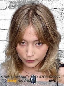 A woman with shoulder-length, layered blonde hair poses in front of a white brick wall